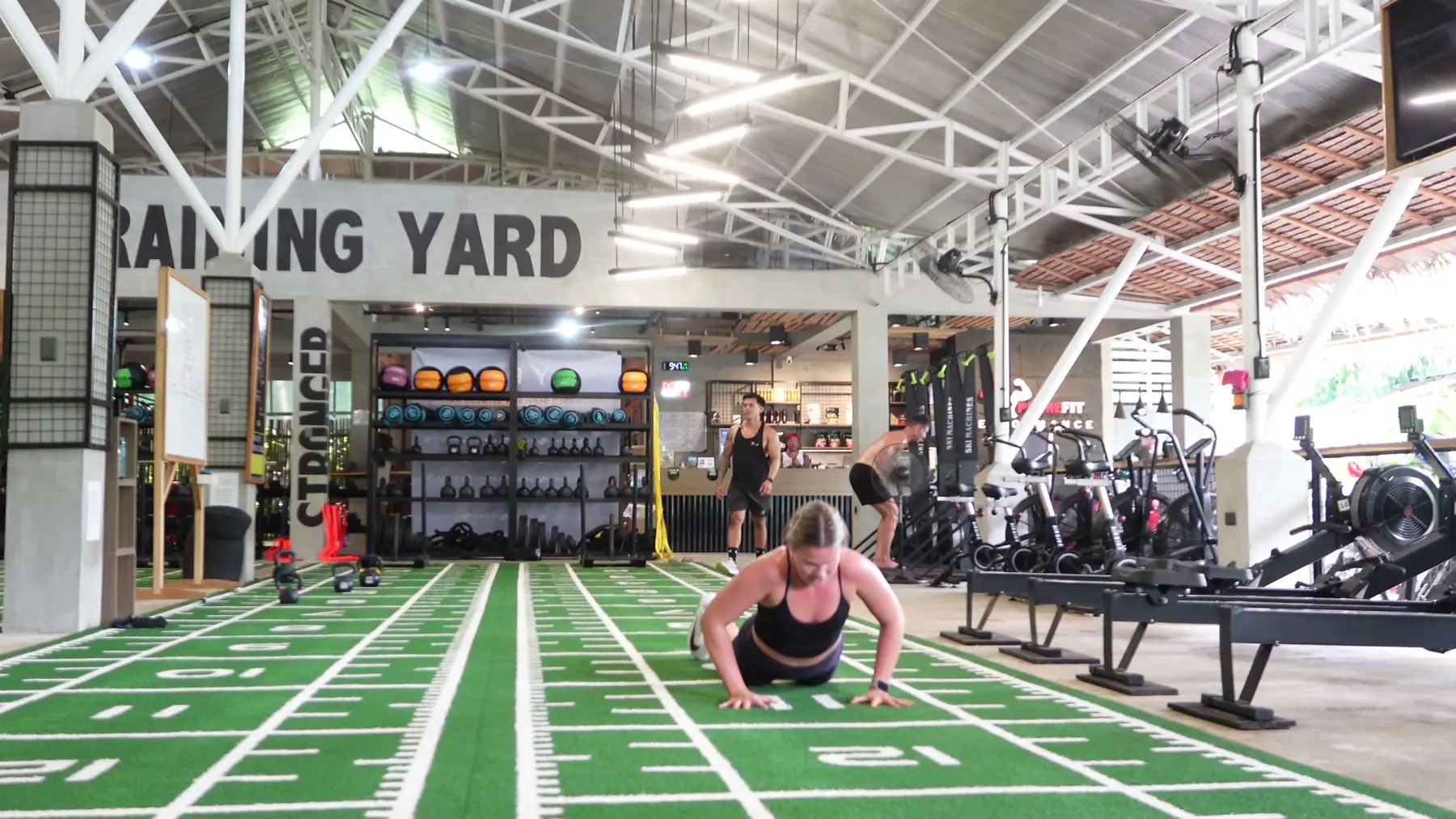 Wide view of a covered gym yard with green turf lanes, racks and weights